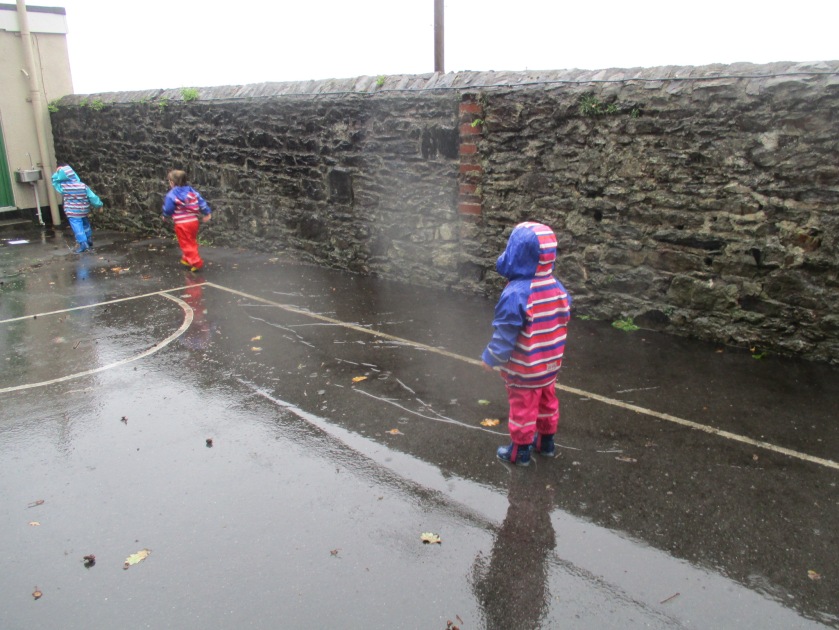 Three children wearing puddle suits playing in puddles in the rain.